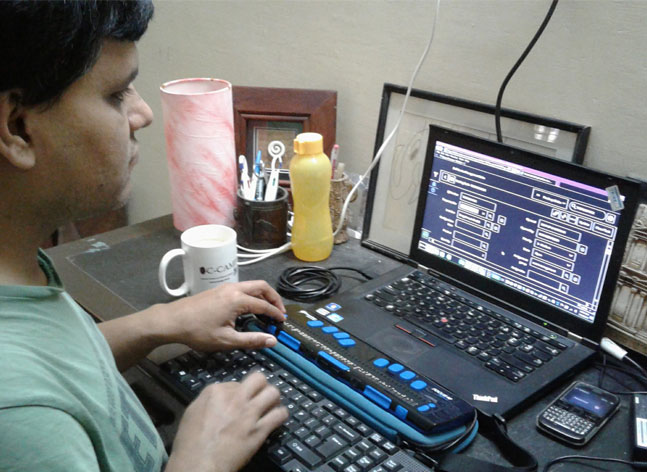 devices Pradip Sinha, a man who is deaf blind sits at a table with one hand on a keyboard and the other on a refreshable braille display. A laptop and cellphone are seen in front of him.