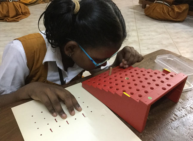 A young girl wearing spectacles leans over a table to place a peg in a pegboard. One hand rests on dots and lines on a sheet beside her.