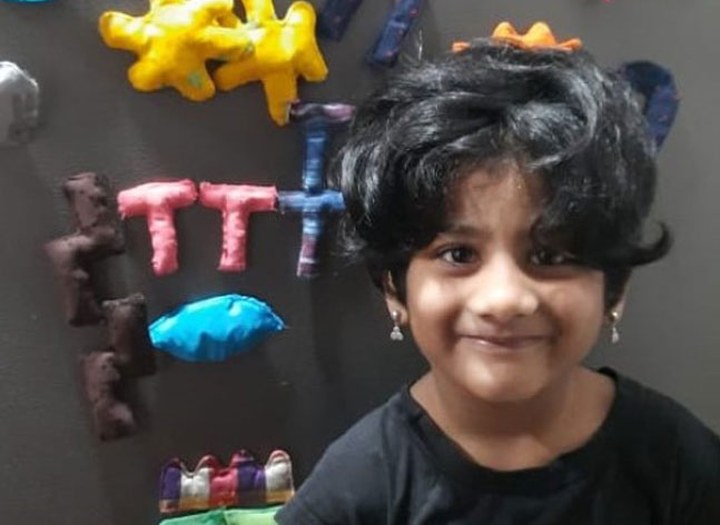 A young girl stands smiling proudly in front of her picture created with cloth covered shapes on the fridge behind her.