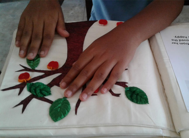 Two small hands explore the tactile image of a tree with leaves and apples in an open book.