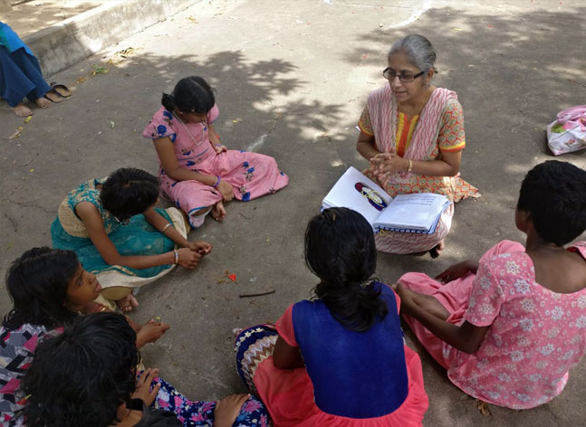 A lady sits on the ground outdoors with a tactile story book open on her lap. A group of girls, their heads bent down, listen to her.